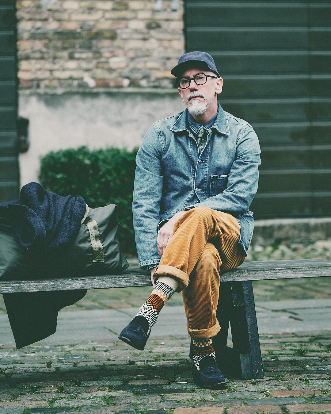 Freddy sitting on a bench in a denim jacket, navy cap and camel corduroy trousers