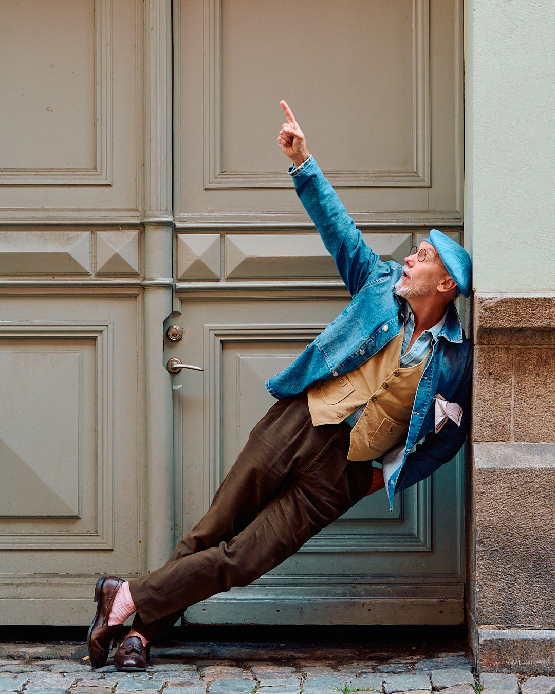 Freddy leaning against an old door, pointing upwards, in a denim jacket and beret