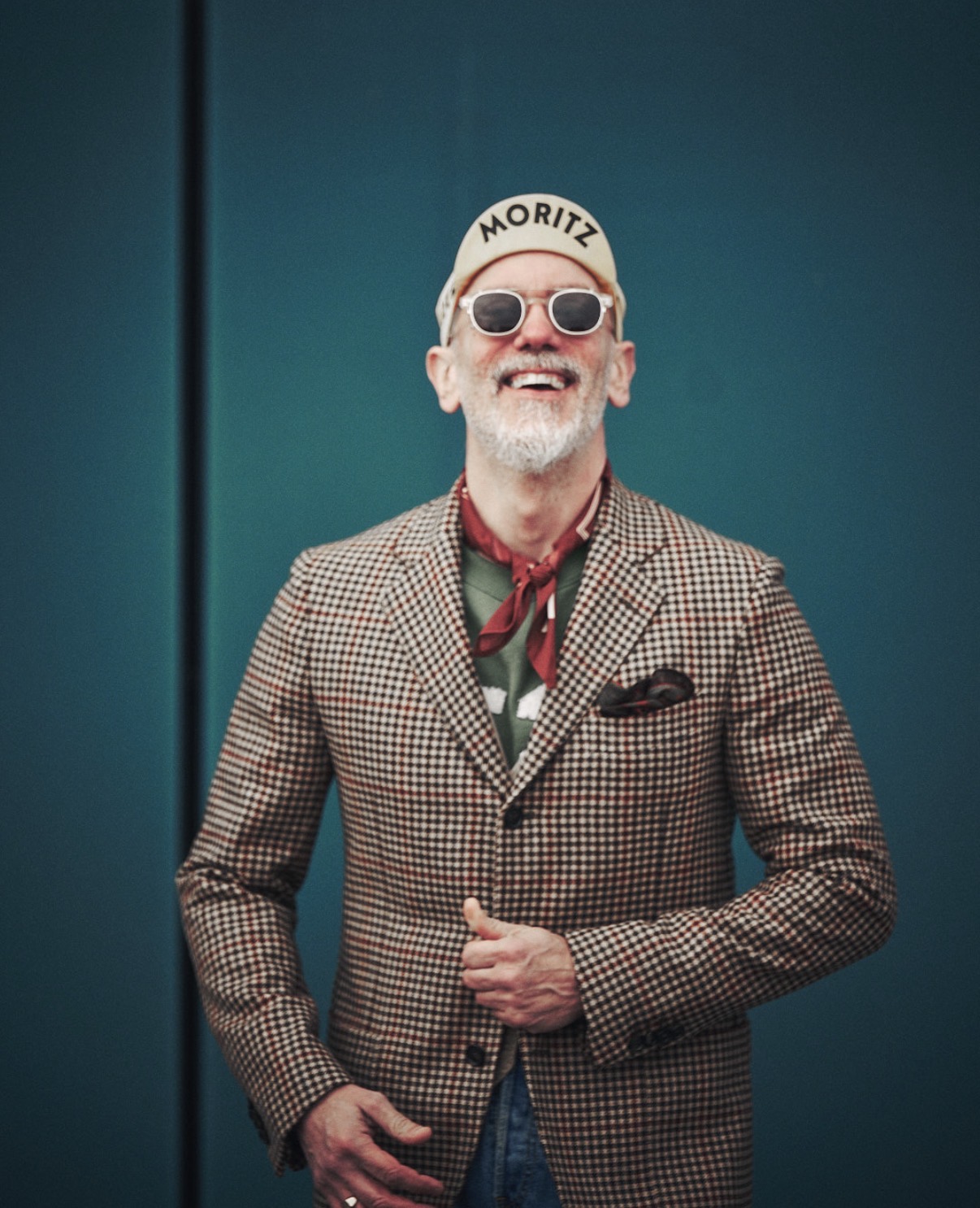 Freddy laughing in a houndstooth jacket and cap against a teal wall
